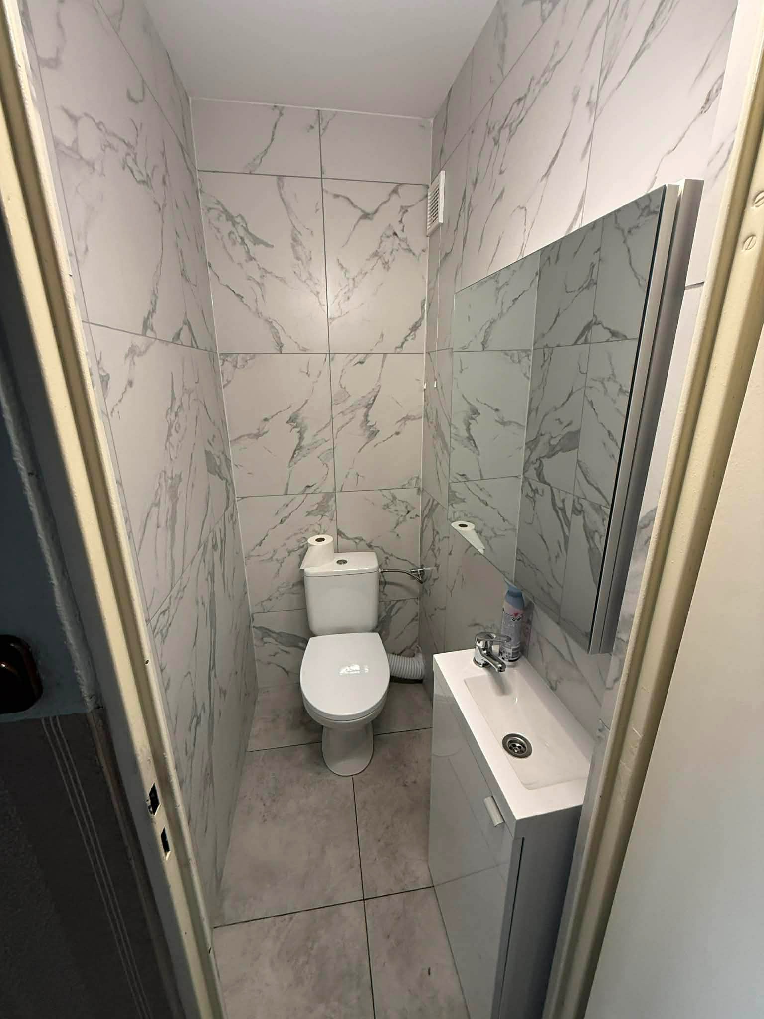 Wąska łazienka po remoncie: biała toaleta, umywalka z szafką i lustro. Ściany wyłożone płytkami imitującymi marmur. Widok zza futryny.