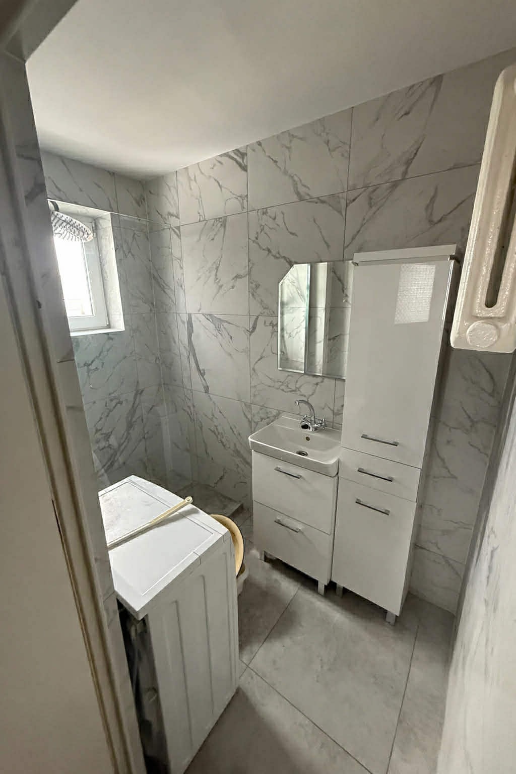 Wykończona łazienka w bieli i szarości z marmurkowymi płytkami. Widoczna umywalka z szafką, lustro, pralka i grzejnik. Minimalistyczny design.