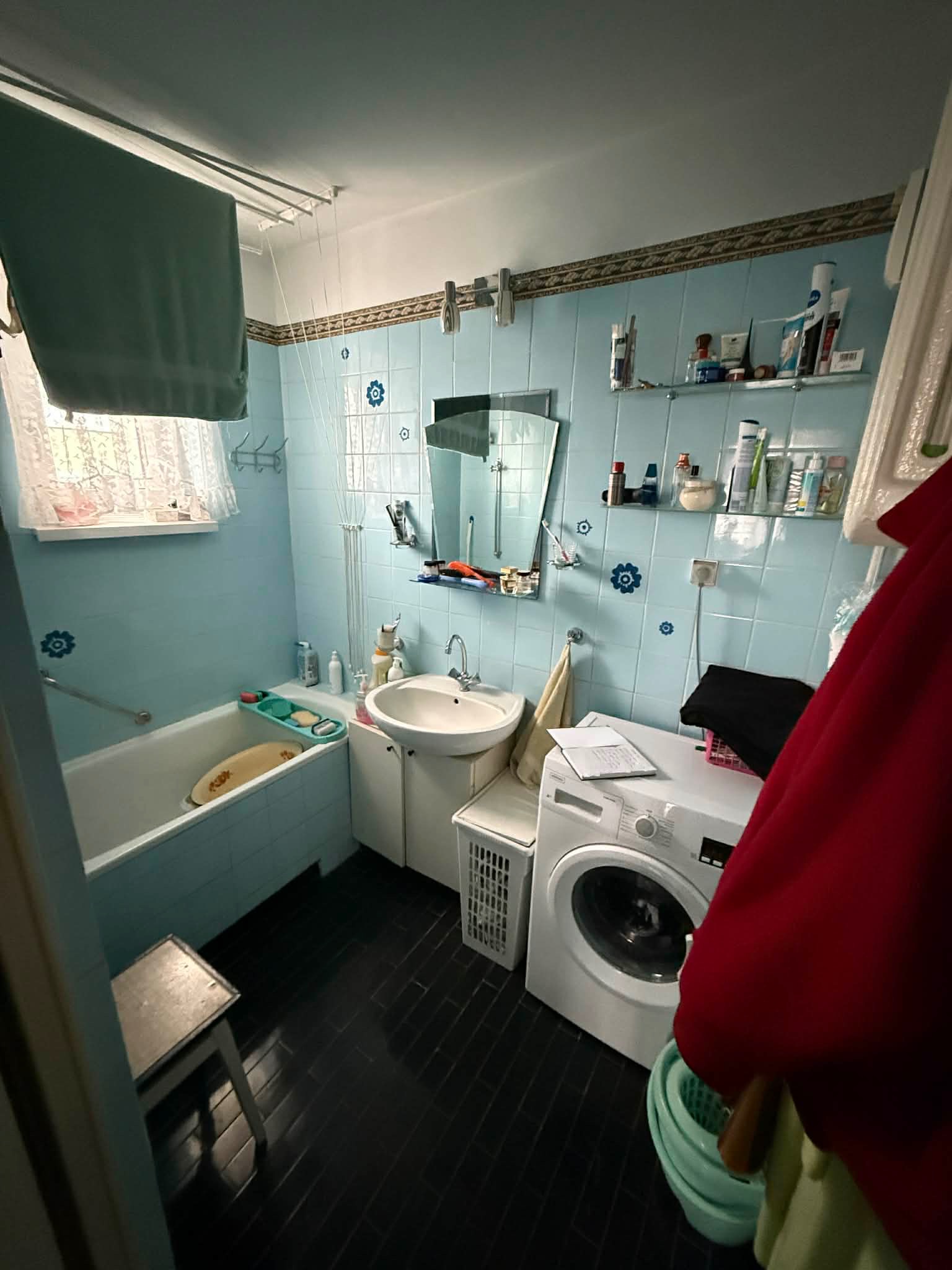 Niebieska łazienka z wanną, umywalką i pralką. Na ścianach płytki z motywem kwiatowym. Widoczny sznurek na pranie i akcesoria łazienkowe na półkach.