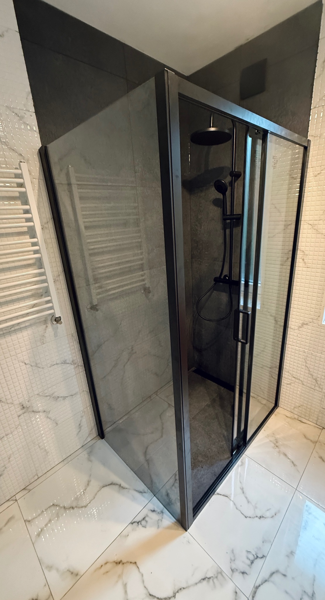 Nowoczesna kabina prysznicowa z czarnymi profilami i deszczownicą, w łazience wykończonej białymi płytkami imitującymi marmur. Widoczny grzejnik drabinkowy.