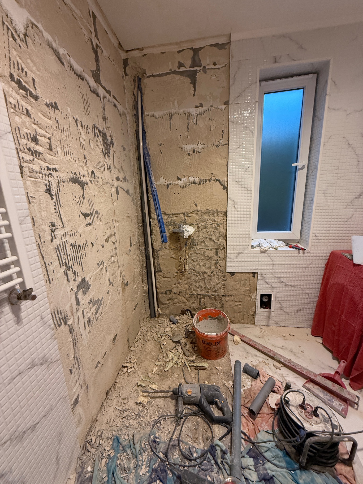 Fragment łazienki w trakcie prac: częściowo położone białe płytki, ściana przygotowana pod płytki, widoczne narzędzia i materiały budowlane. Okno z niebieskim światłem.