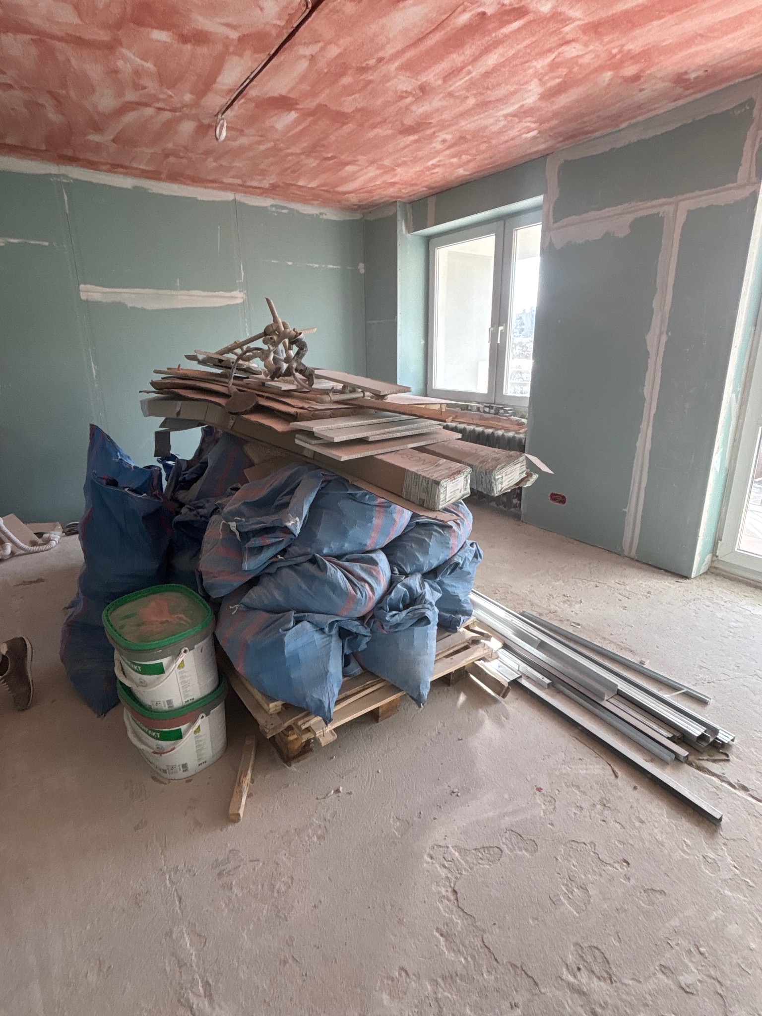 Wnętrze w trakcie remontu: worki z gruzem, płyty gipsowe, profile metalowe i puszki z farbą na tle surowych ścian i sufitu. Widoczne okno i fragment podłogi.