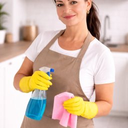 VAN GLOBAL SPÓŁKA Z OGRANICZONĄ ODPOWIEDZIALNOŚCIĄ - Uśmiechnięta kobieta w fartuchu i żółtych rękawicach trzymająca butelkę ze środkiem czyszczącym i różową ściereczkę na tle jasnej kuchni.