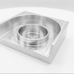 SON - MET PATRYK MORAWSKI - Precyzyjnie obrobiony element z metalu o kwadratowej podstawie i okrągłym wgłębieniem, widoczne ślady obróbki skrawaniem, na białym tle.