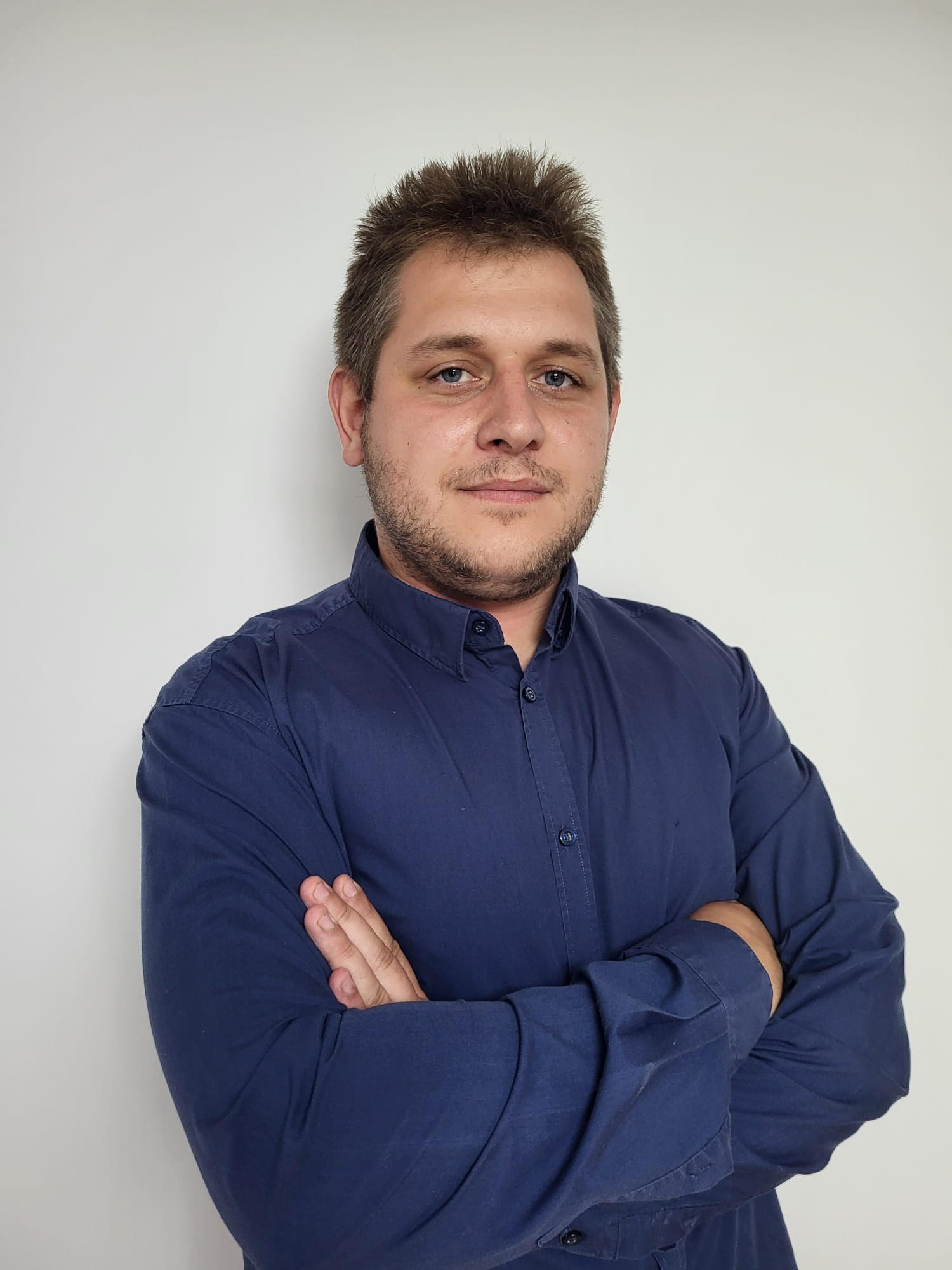 Portret mężczyzny w niebieskiej koszuli z założonymi rękami na tle jasnej ściany. Widoczne zmarszczki mimiczne i zarost. Ujęcie od przodu, skupione na twarzy.