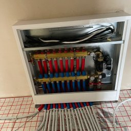 Kacper - Rozdzielacz ogrzewania podłogowego w białej szafce, z widocznymi rurami w kolorach czerwonym i niebieskim, ułożonymi na izolacji podłogowej z siatką.