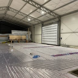 Kacper - Rozległa instalacja ogrzewania podłogowego w hali, rury ułożone na siatce, widoczne narzędzia i brama w tle. Jasne oświetlenie, surowy industrialny charakter.