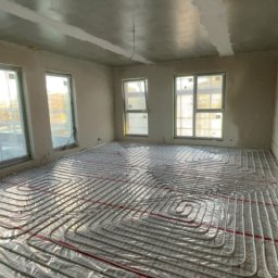 Kacper - Ułożona instalacja ogrzewania podłogowego w surowym wnętrzu z trzema oknami. Rury grzewcze rozłożone spiralnie na folii z czerwoną siatką, przygotowane do zalania posadzką.