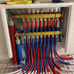 Kacper - Rozdzielacz ogrzewania podłogowego z rurami w kolorach czerwonym i niebieskim, zamontowany w białej szafce, widoczne narzędzia. Instalacja na folii grzewczej.