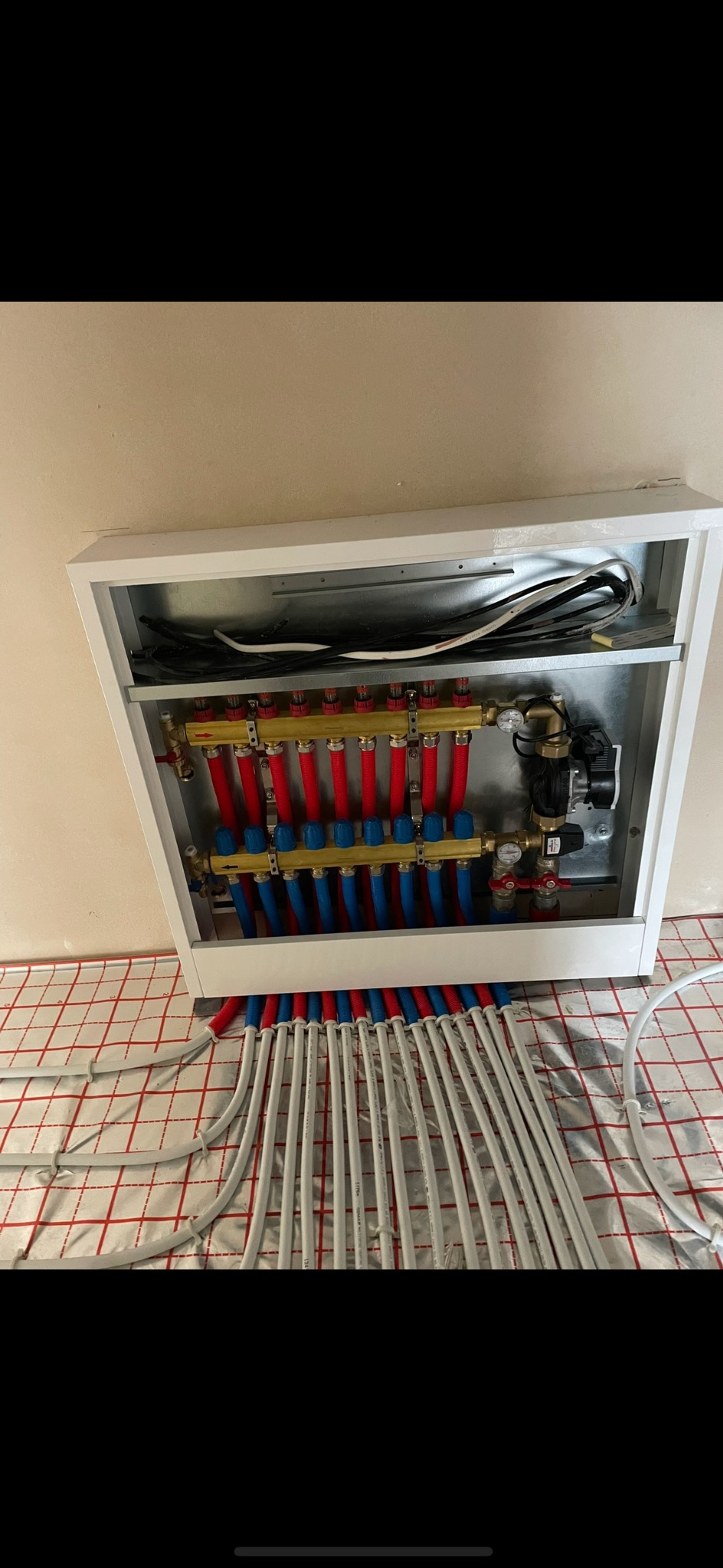 Rozdzielacz ogrzewania podłogowego w białej szafce, z widocznymi rurami w kolorach czerwonym i niebieskim, ułożonymi na izolacji podłogowej z siatką.
