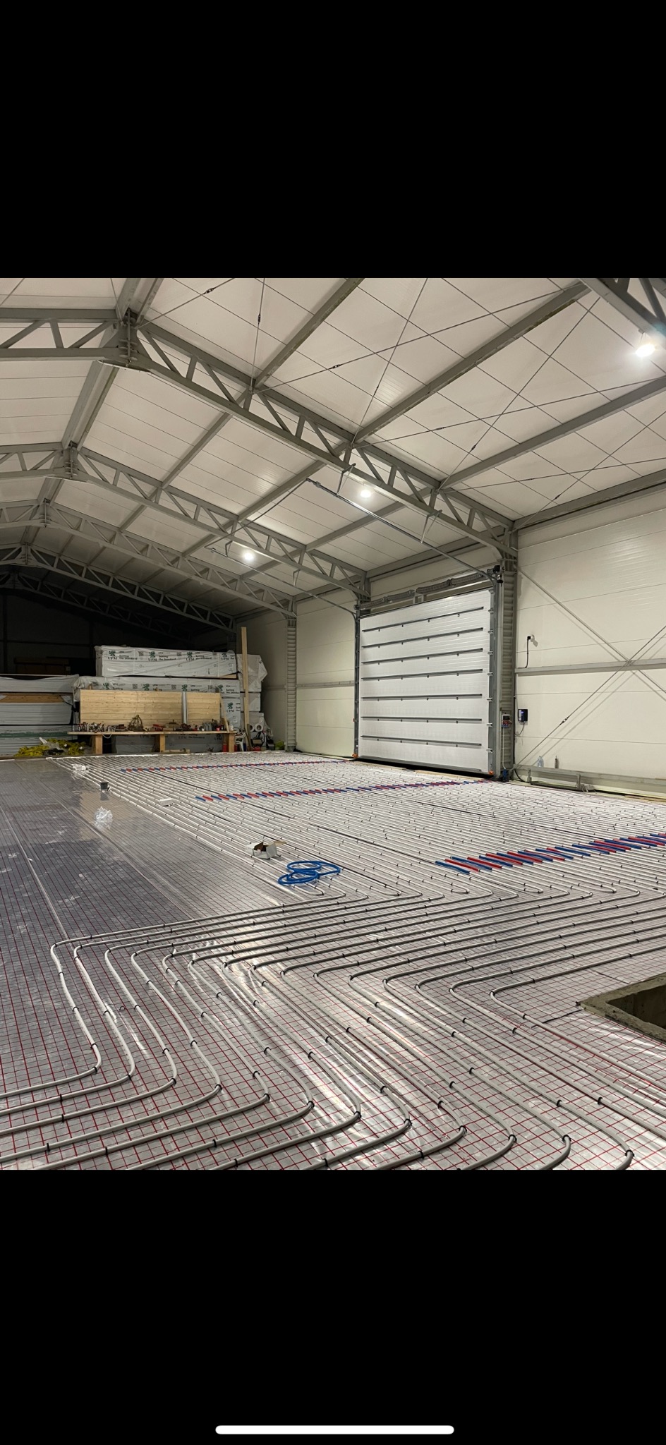 Rozległa instalacja ogrzewania podłogowego w hali, rury ułożone na siatce, widoczne narzędzia i brama w tle. Jasne oświetlenie, surowy industrialny charakter.