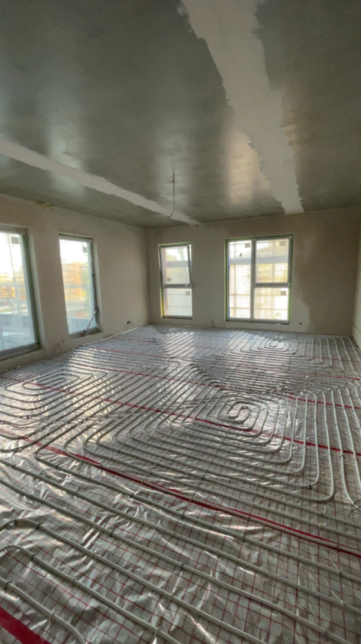 Ułożona instalacja ogrzewania podłogowego w surowym wnętrzu z trzema oknami. Rury grzewcze rozłożone spiralnie na folii z czerwoną siatką, przygotowane do zalania posadzką.