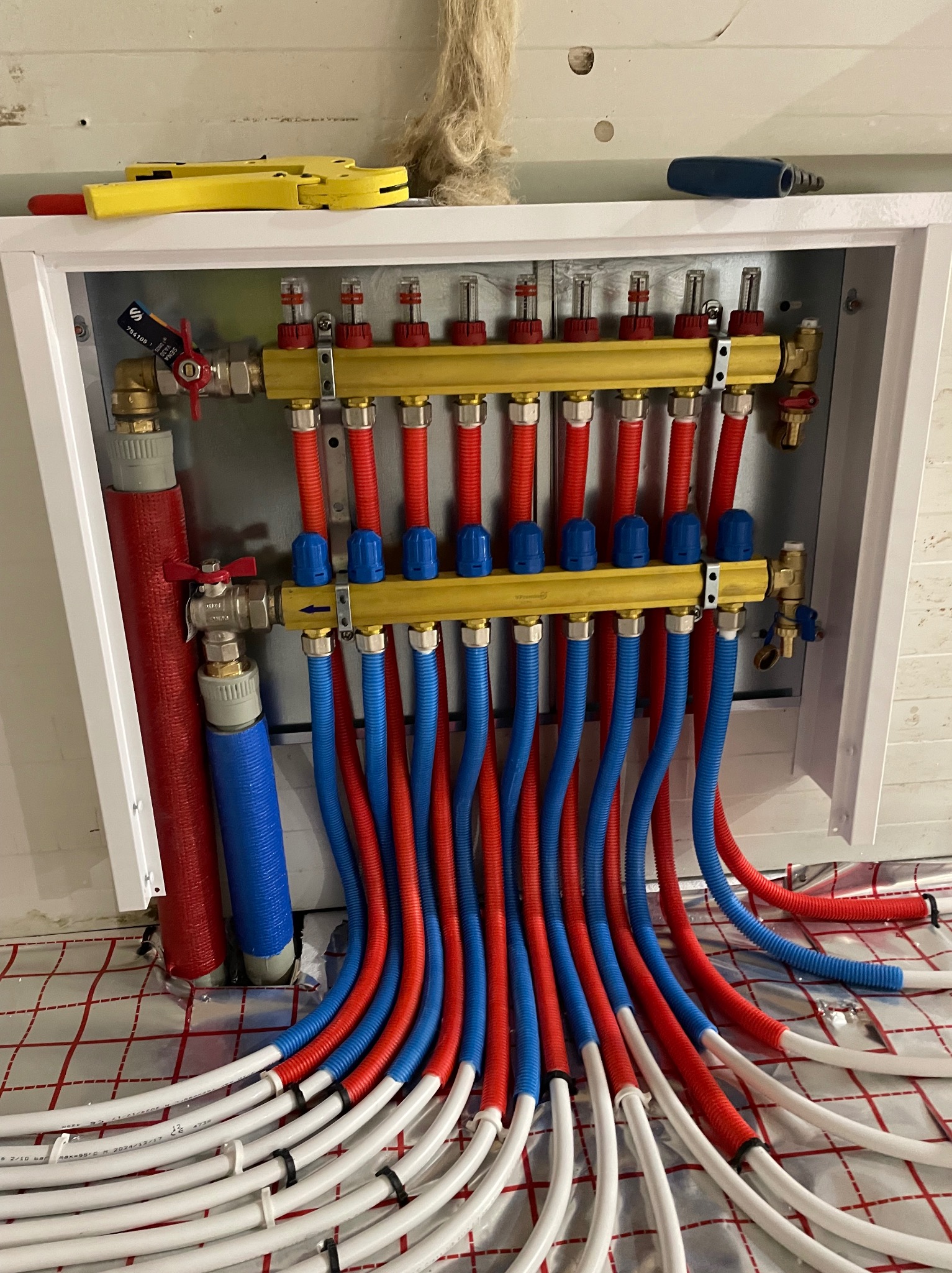 Rozdzielacz ogrzewania podłogowego z rurami w kolorach czerwonym i niebieskim, zamontowany w białej szafce, widoczne narzędzia. Instalacja na folii grzewczej.