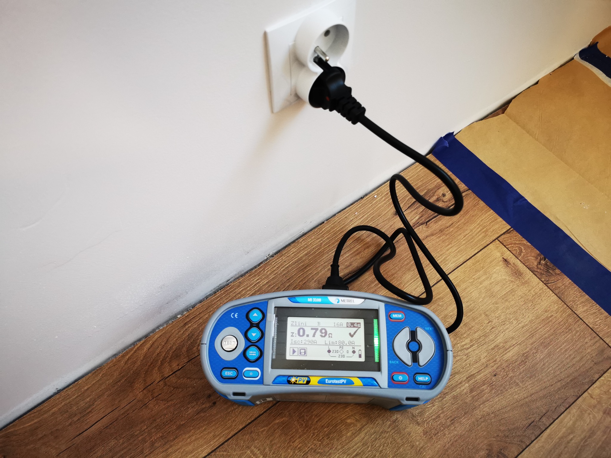 Miernik Metrel MI 3108 podłączony do gniazdka elektrycznego, wyświetlający wynik pomiaru impedancji pętli zwarcia na drewnianej podłodze, zabezpieczonej kartonem.