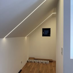 Usługi Elektryczne Maksymilian Lis - Podświetlone ledami poddasze ze skośnym sufitem i małym oknem. Na podłodze panele i poziomica. Nowoczesne oświetlenie wkomponowane w architekturę wnętrza.