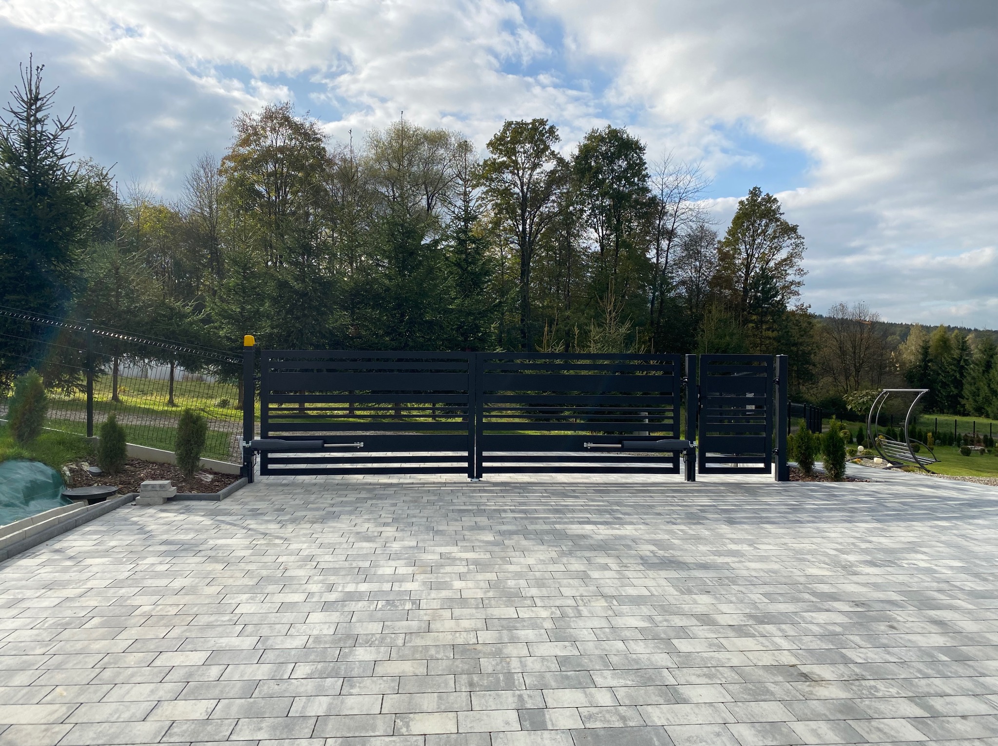 Nowoczesna, przesuwna brama wjazdowa w kolorze antracytowym, z widocznym mechanizmem automatyki, na tle brukowanej posesji i zieleni drzew.