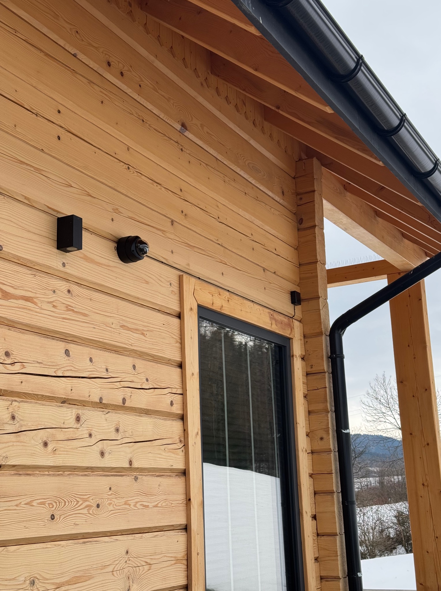 Kamera monitoringu i czujnik na drewnianej ścianie domu z oknem. Widoczny fragment dachu z rynną i krajobraz w tle. Zewnętrzny montaż systemu zabezpieczeń.