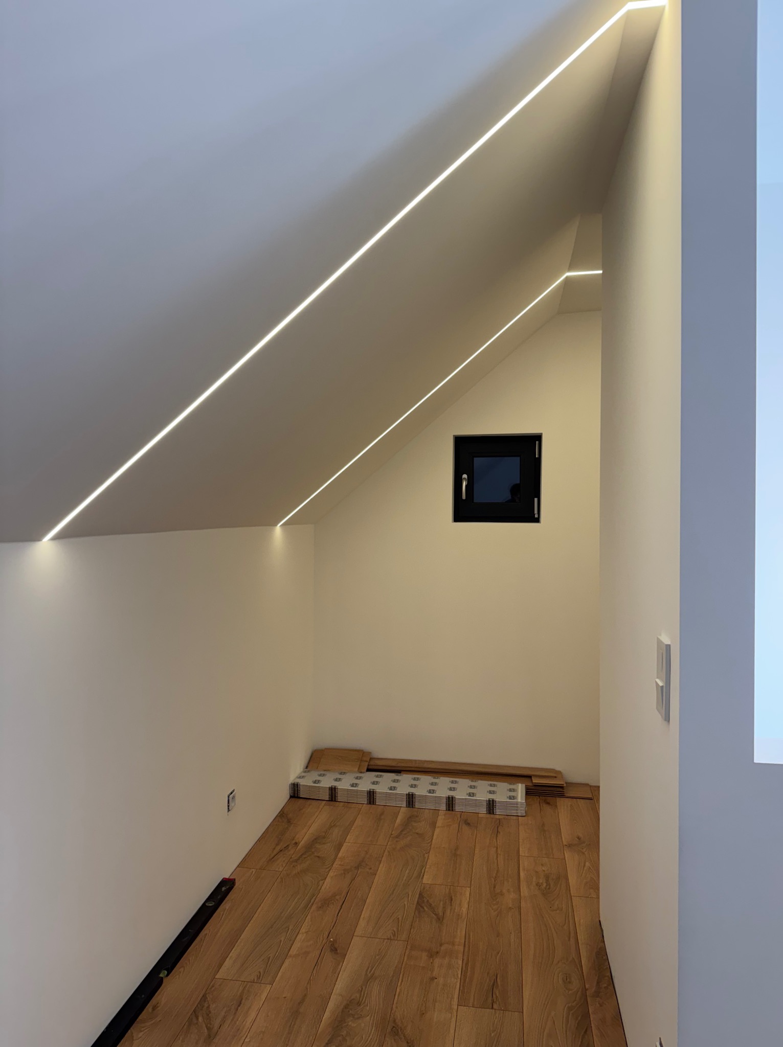 Podświetlone ledami poddasze ze skośnym sufitem i małym oknem. Na podłodze panele i poziomica. Nowoczesne oświetlenie wkomponowane w architekturę wnętrza.