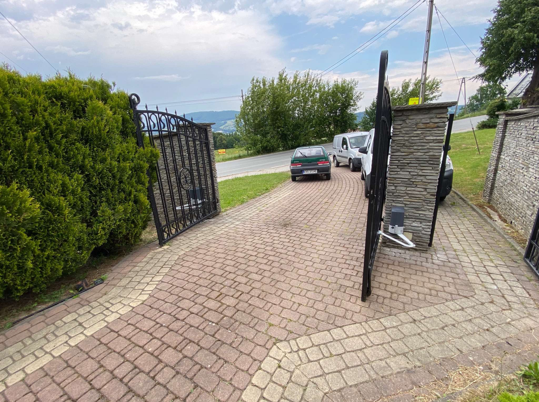 Otwarta, kuta brama wjazdowa z automatyką, prowadząca na posesję z brukowanym podjazdem. W tle widoczne samochody i zieleń.