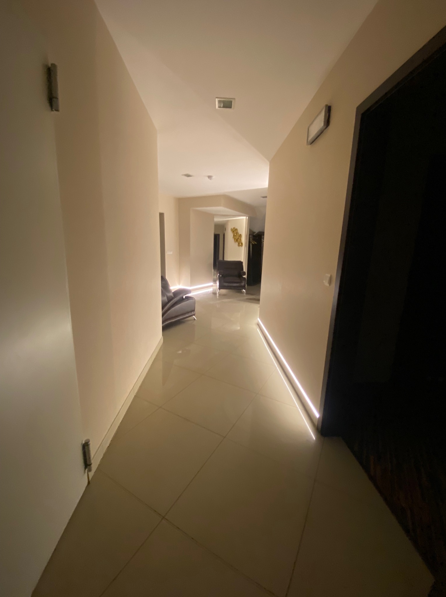 Długi korytarz z beżowymi ścianami i podświetleniem LED przy podłodze, prowadzący do salonu z szarą sofą. Minimalistyczny design, ciepłe oświetlenie.