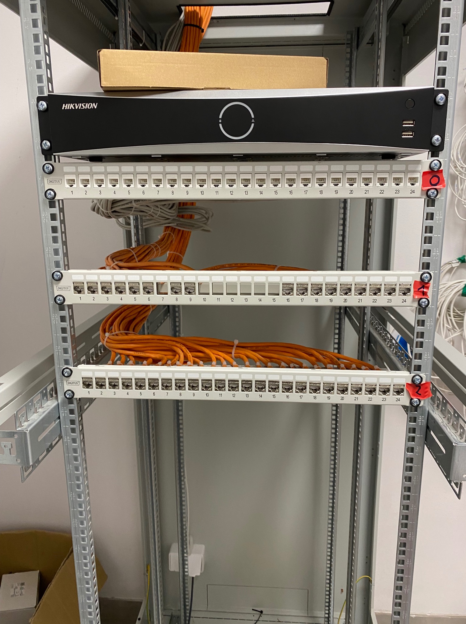 Panel krosowniczy Digitus w szafie rack z pomarańczowymi kablami sieciowymi. Widoczny rejestrator Hikvision. Instalacja sieci komputerowej w serwerowni.