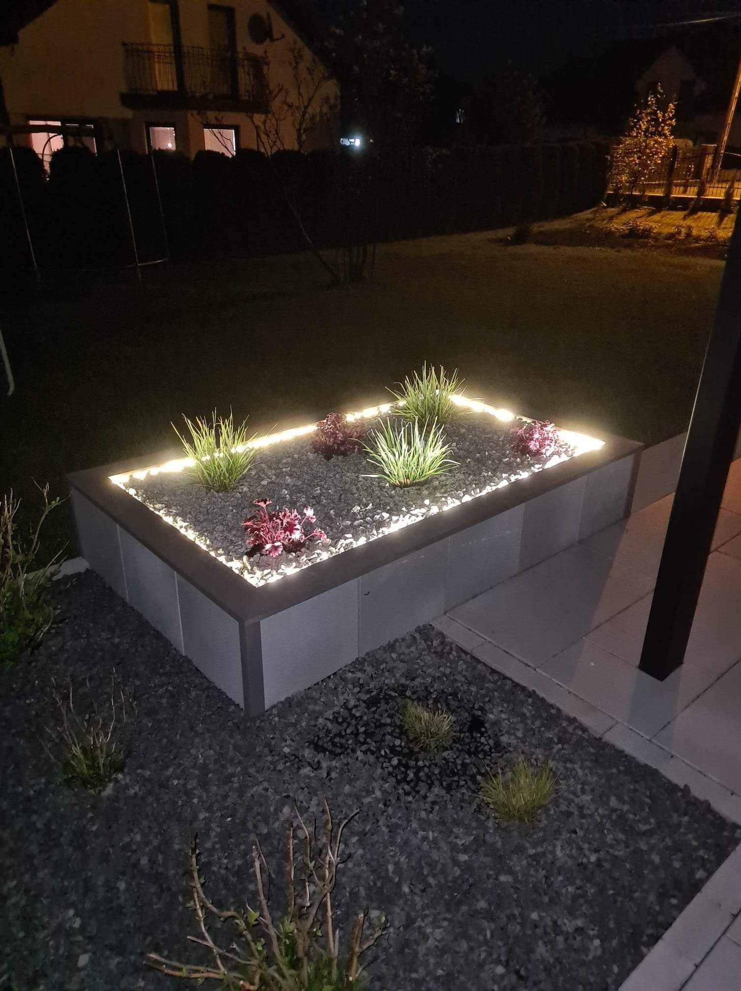 Podświetlona rabata z roślinami i kamieniami w ogrodzie nocą. Dekoracyjne oświetlenie LED podkreśla kształt rabaty i dodaje uroku otoczeniu domu.