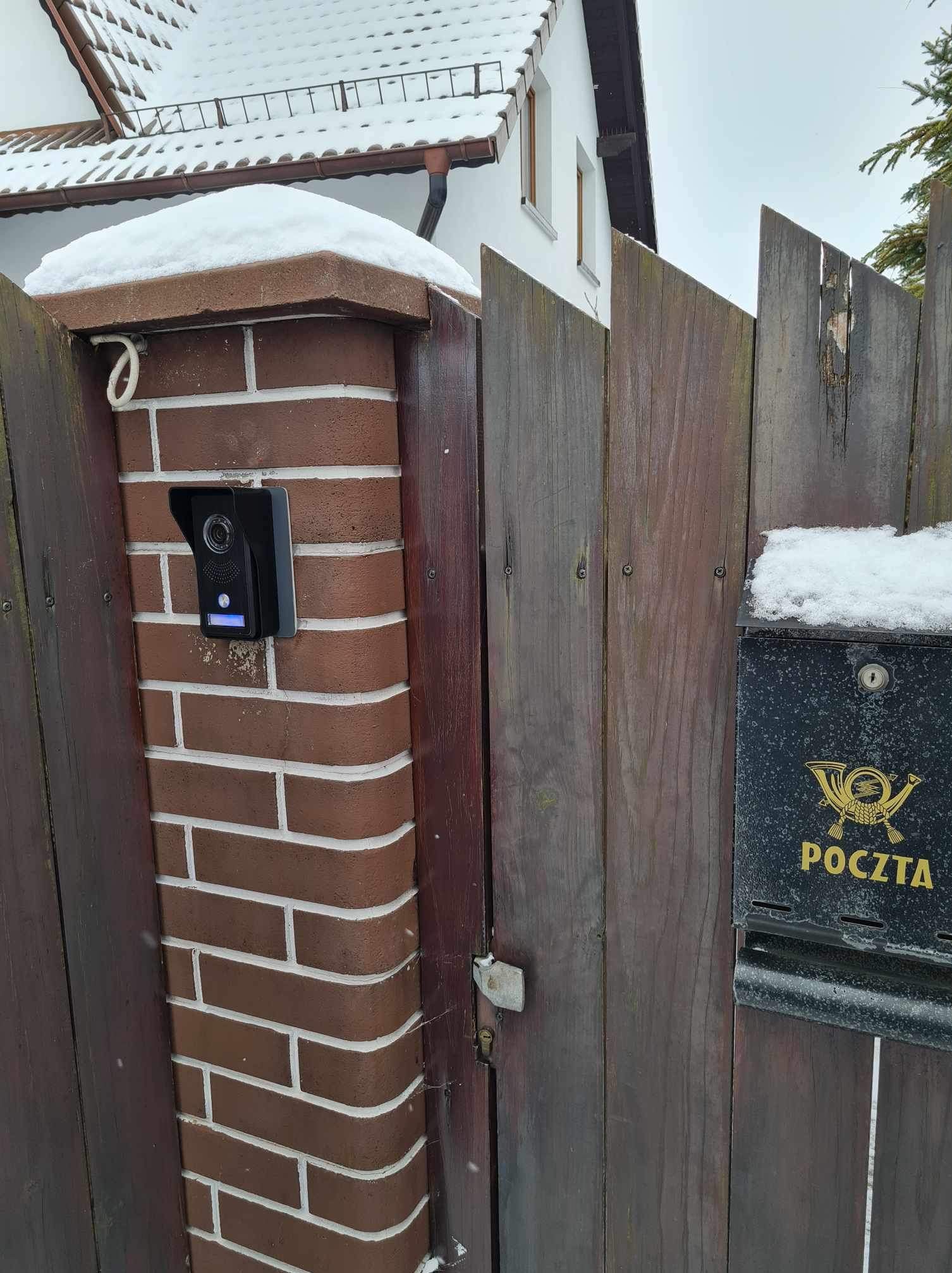 Nowoczesny wideodomofon zamontowany na ceglanym słupku ogrodzenia, obok drewniana furtka i skrzynka pocztowa w zimowej scenerii.