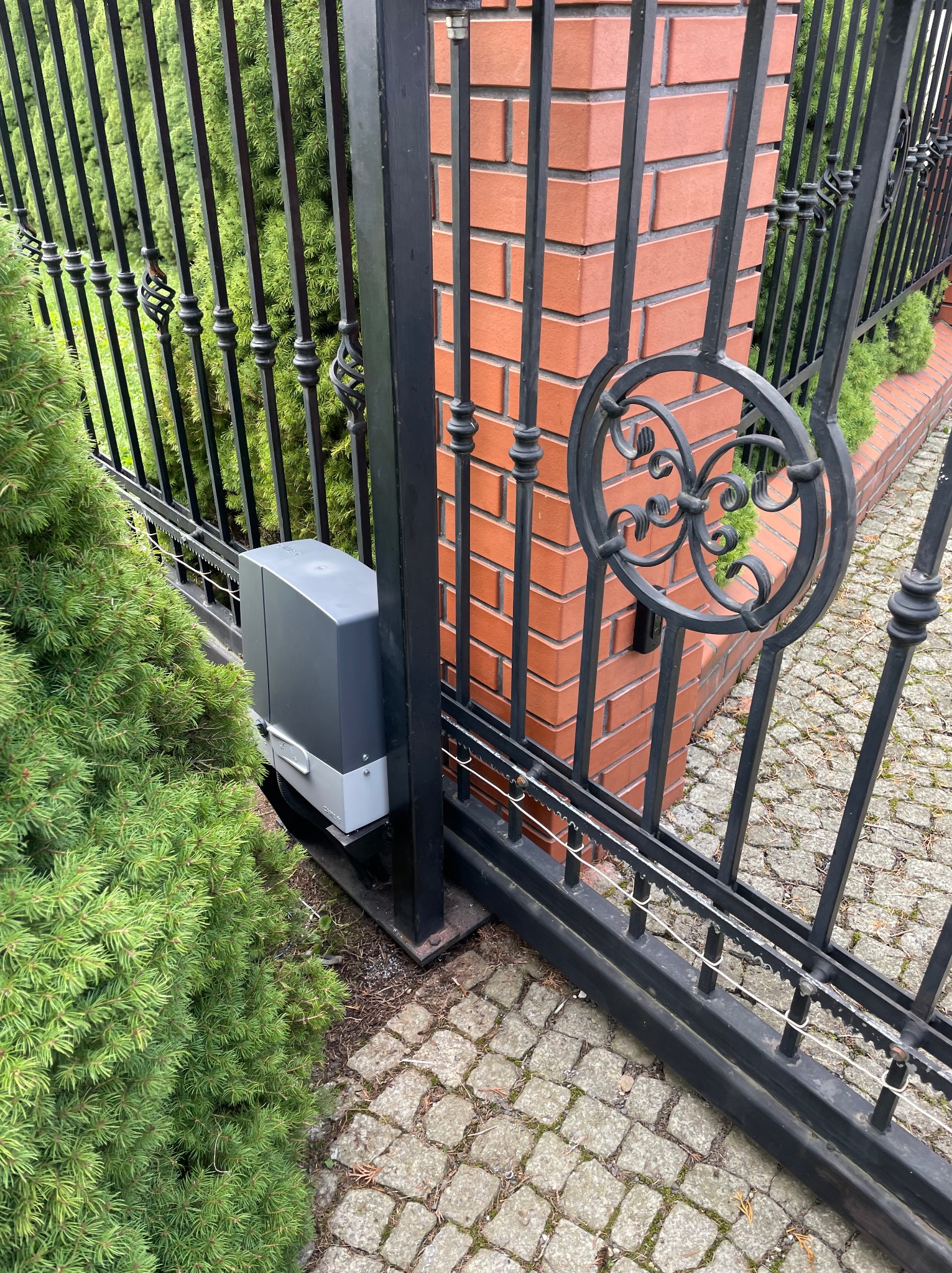 Czarna, metalowa brama wjazdowa z automatyką, widoczny napęd bramy. Brama zdobiona ornamentami, obok rośnie iglak, podłoże wyłożone kostką brukową.