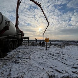 SIMS HOUSE SP. Z O. O. - Zimowy krajobraz budowy: betonowóz z wysięgnikiem pompuje beton do szalunków fundamentowych. W tle pochmurne niebo i sylwetki pracowników.