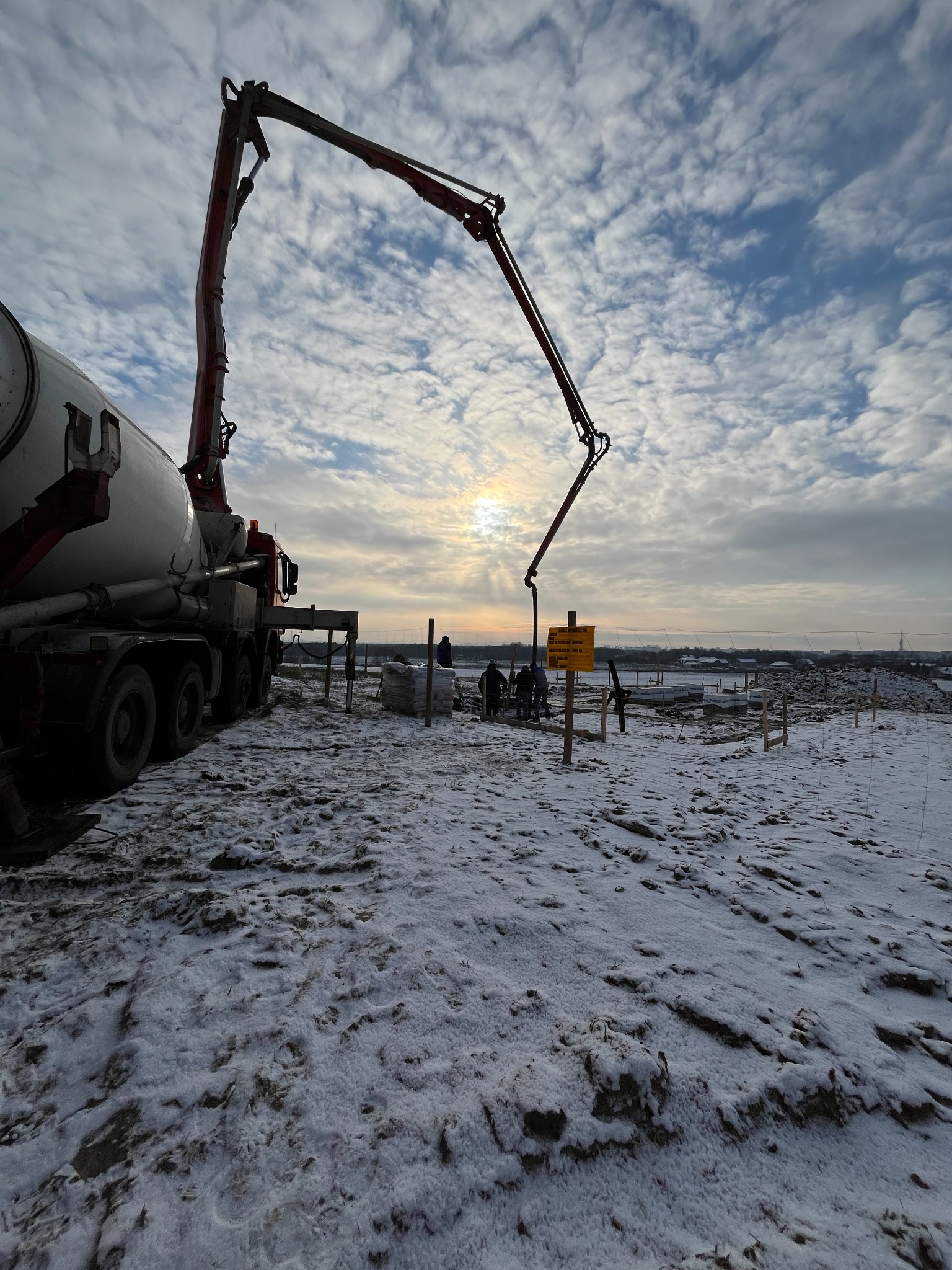Zimowy krajobraz budowy: betonowóz z wysięgnikiem pompuje beton do szalunków fundamentowych. W tle pochmurne niebo i sylwetki pracowników.