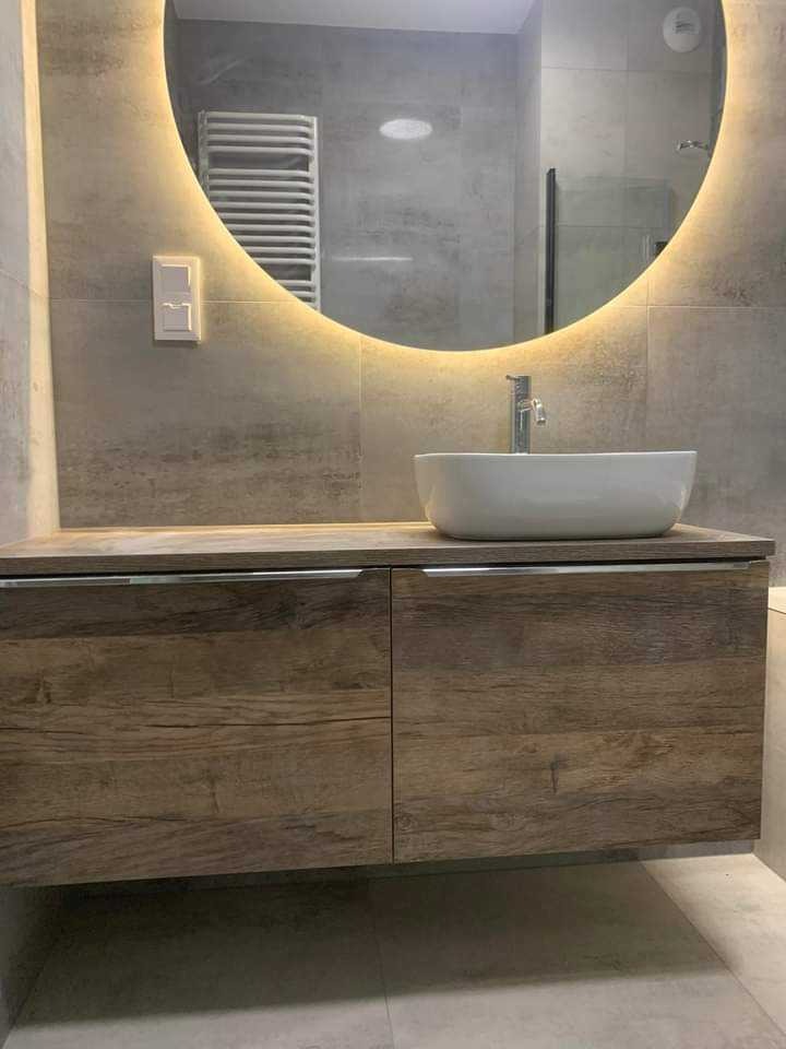 Nowoczesna łazienka z okrągłym lustrem podświetlanym LED, umywalką nablatową i szafką z drewnopodobnym frontem. Minimalistyczny design, szare płytki.