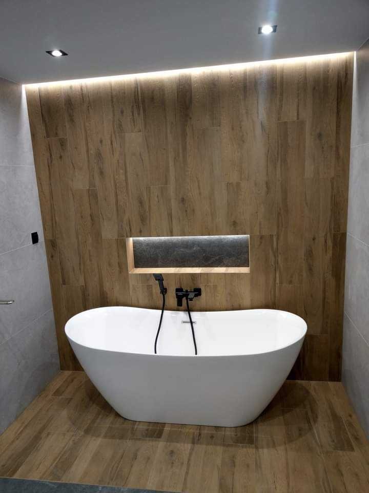 Nowoczesna łazienka z wolnostojącą białą wanną na tle ściany wyłożonej płytkami imitującymi drewno, podświetlona listwą LED. Minimalistyczny design.