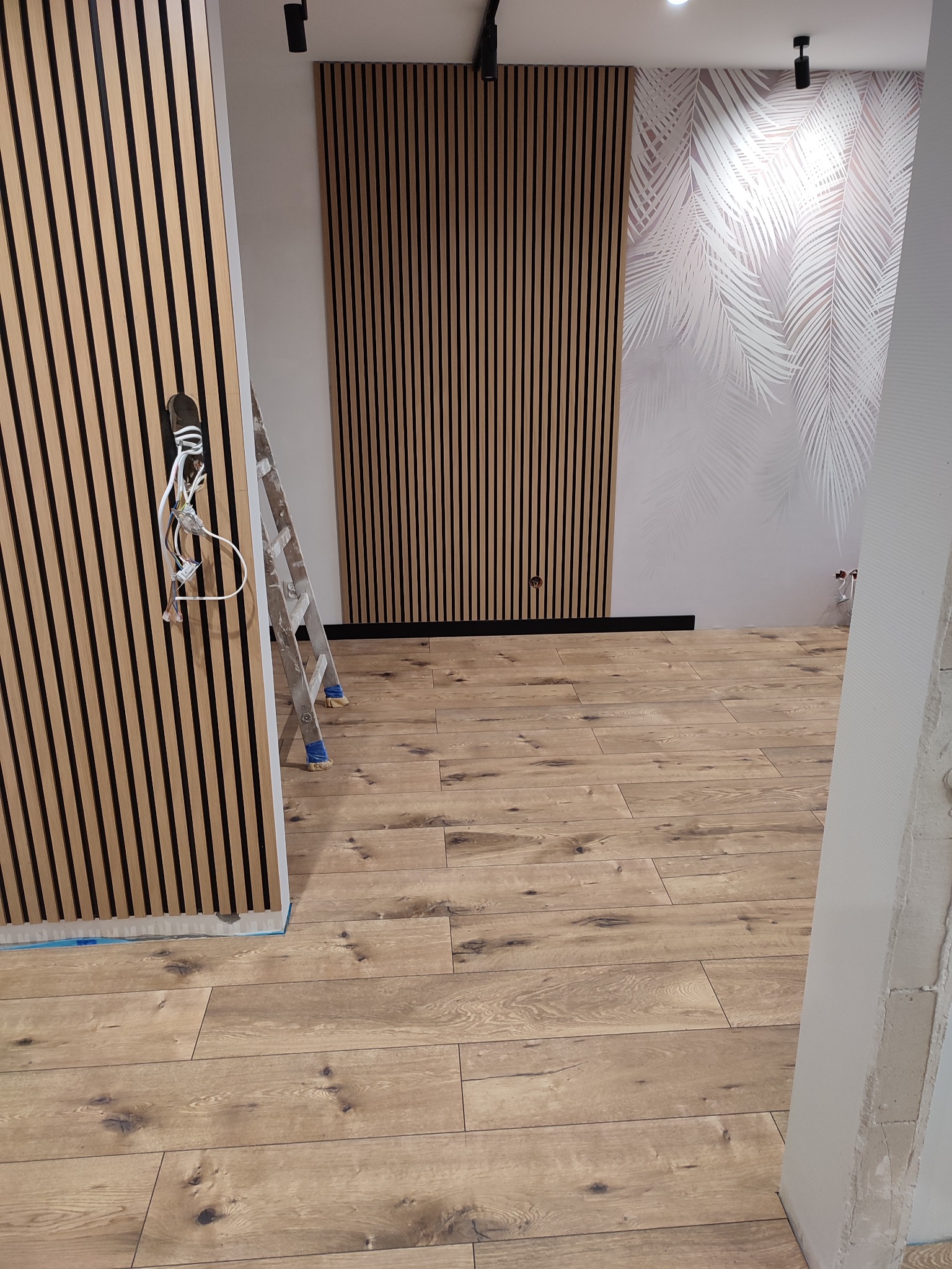 Wykończone wnętrze z drewnianymi lamelami na ścianach, podłogą z paneli imitujących drewno i tapetą w liście. Widoczna drabina i instalacja elektryczna.