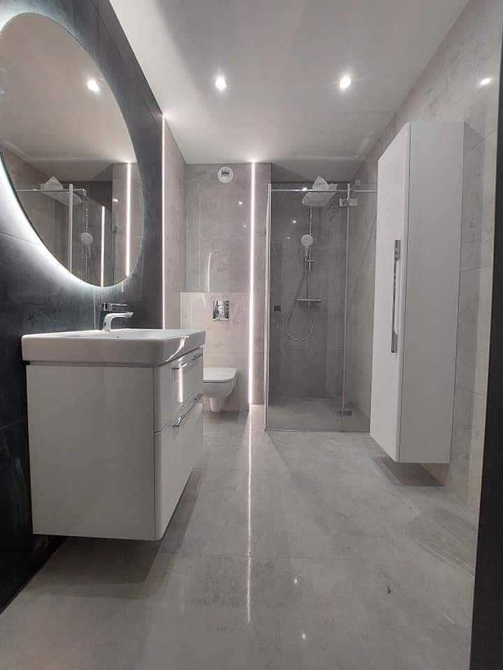 Nowoczesna łazienka w szarościach z owalnym lustrem podświetlanym LED, kabiną prysznicową typu walk-in i białymi meblami łazienkowymi. Minimalistyczny design.