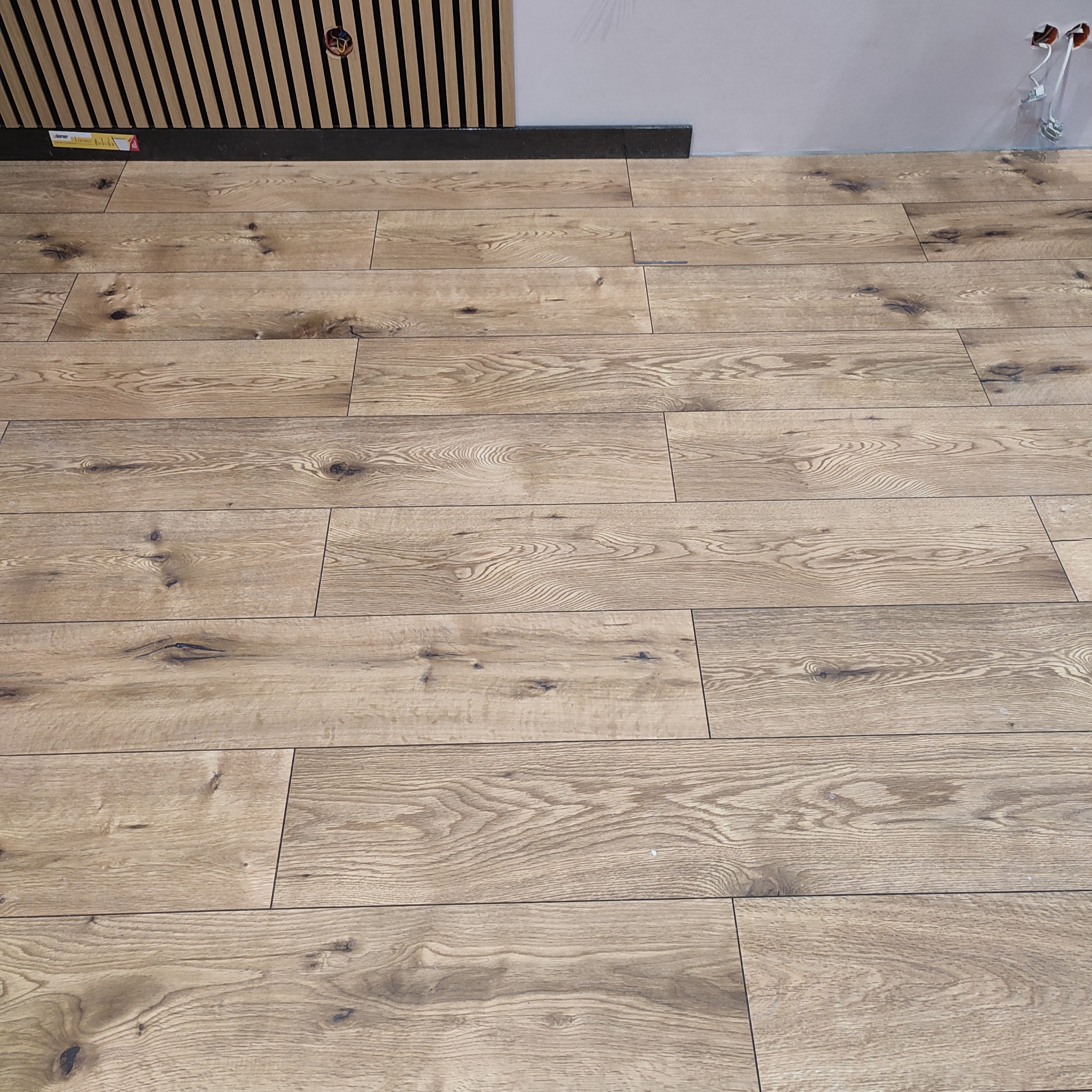 Ułożone panele podłogowe w odcieniach brązu, widoczne fragmenty ściany z listwami dekoracyjnymi i instalacją elektryczną. Układanie paneli.