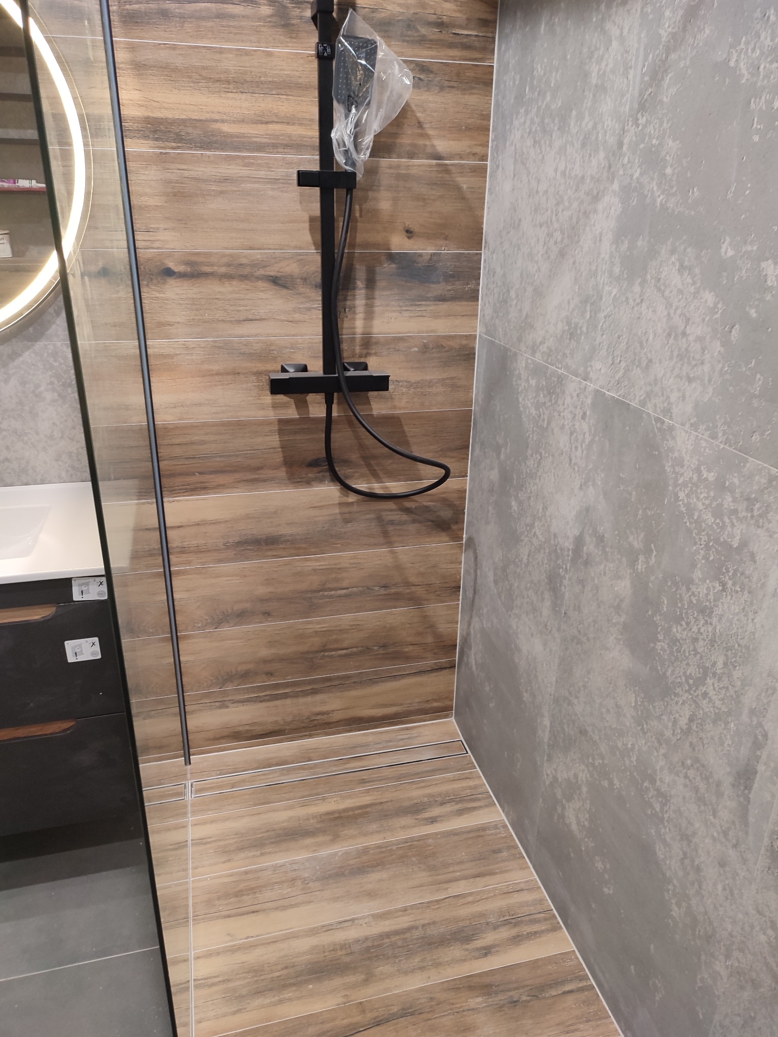 Nowoczesna kabina prysznicowa z drewnopodobnymi i szarymi płytkami, czarną armaturą i odpływem liniowym. Widoczny fragment umywalki z szafką.