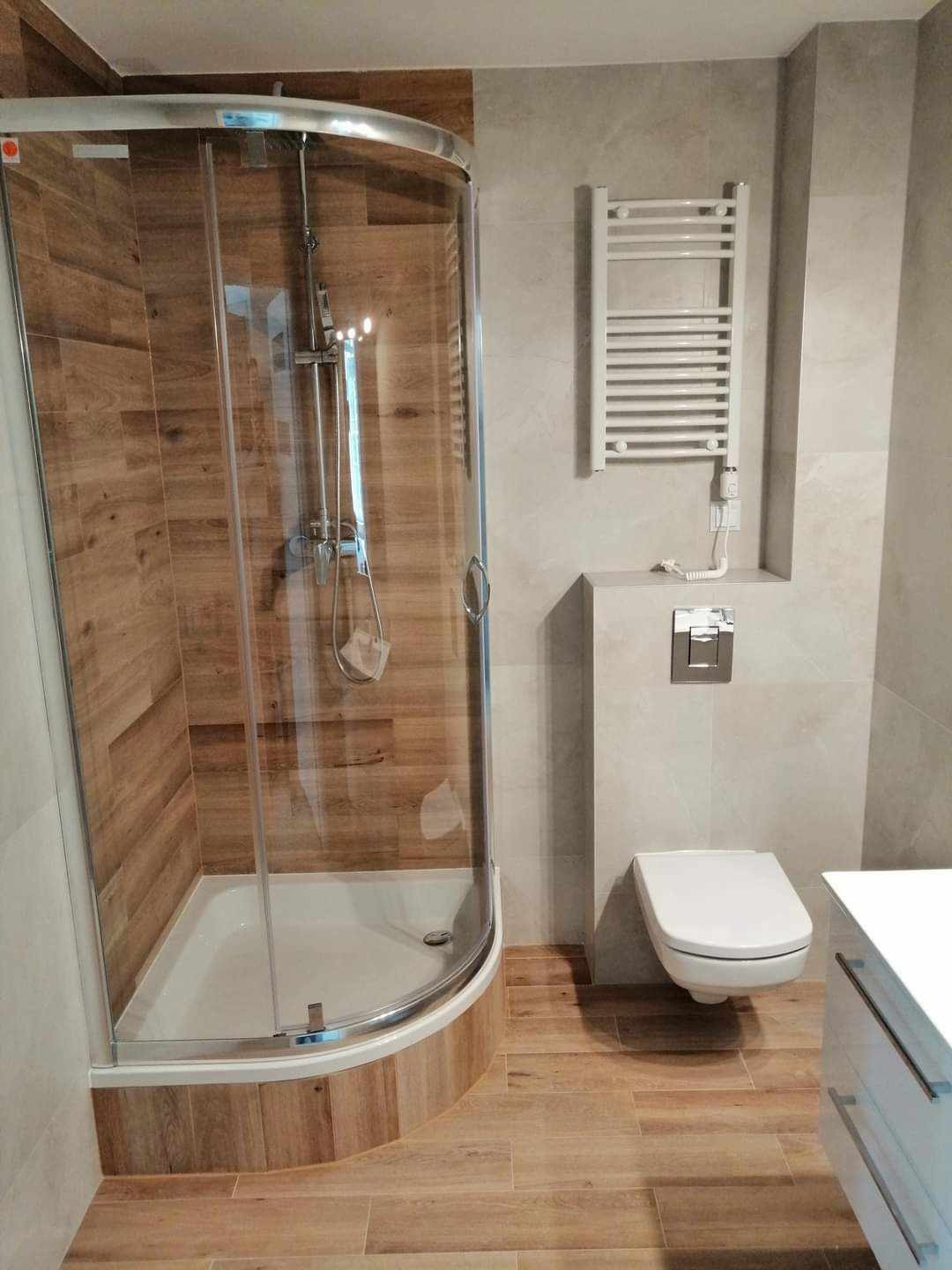 Nowoczesna łazienka z kabiną prysznicową typu walk-in, drewnopodobnymi płytkami i grzejnikiem drabinkowym. Uwagę zwraca minimalistyczna toaleta podwieszana.