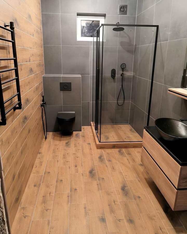 Nowoczesna łazienka z kabiną prysznicową typu walk-in, drewnopodobnymi płytkami na podłodze i ścianie, szarą glazurą i czarną armaturą.