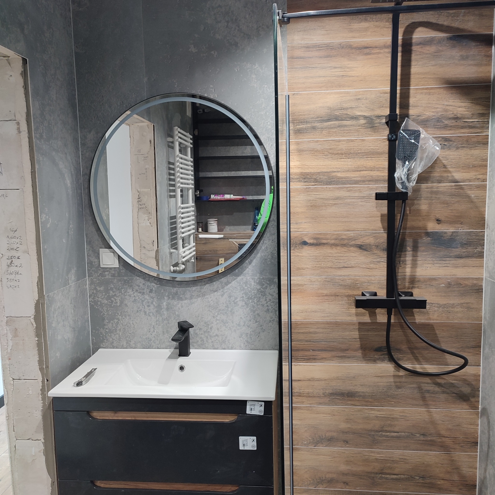 Nowoczesna łazienka z szarą płytką imitującą beton, okrągłe lustro z podświetleniem, czarna armatura i kabina prysznicowa z drewnopodobnymi płytkami.