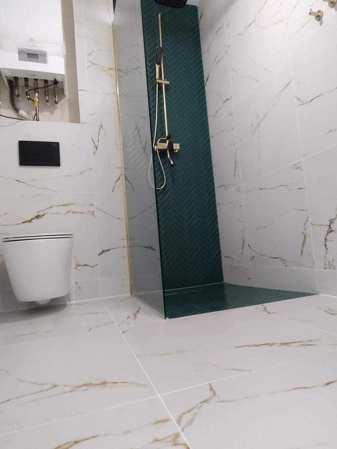 Nowoczesna łazienka z kabiną typu walk-in, wykończona białymi płytkami imitującymi marmur z akcentami złota i zieleni. Widoczny wiszący sedes i złota armatura.