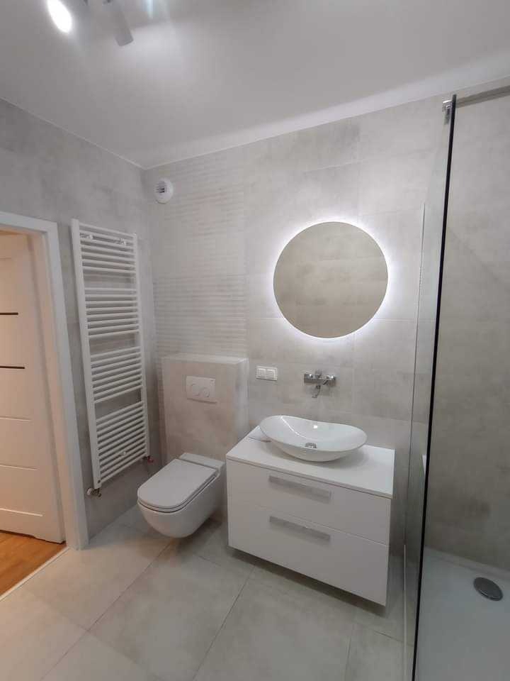 Jasna łazienka z okrągłym lustrem LED, umywalką nablatową na białej szafce i grzejnikiem drabinkowym. Minimalistyczny design, szare płytki i kabina prysznicowa.
