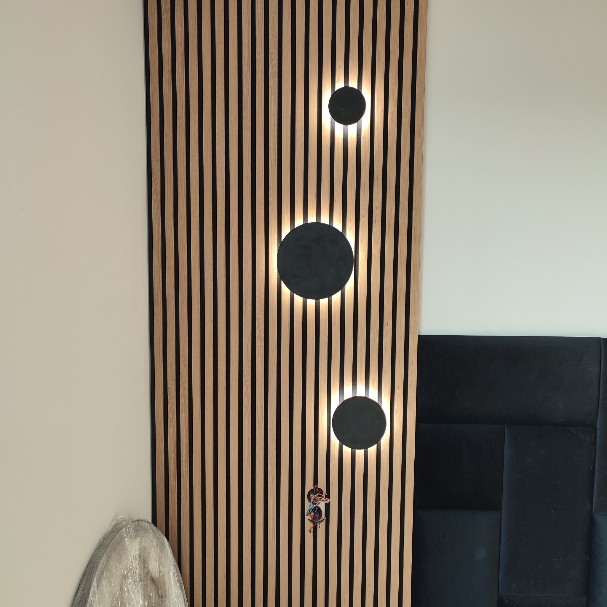 Panel ścienny z drewnianych listew z trzema okrągłymi lampami, widoczne kable elektryczne. Część wezgłowia łóżka i mebel w folii ochronnej.