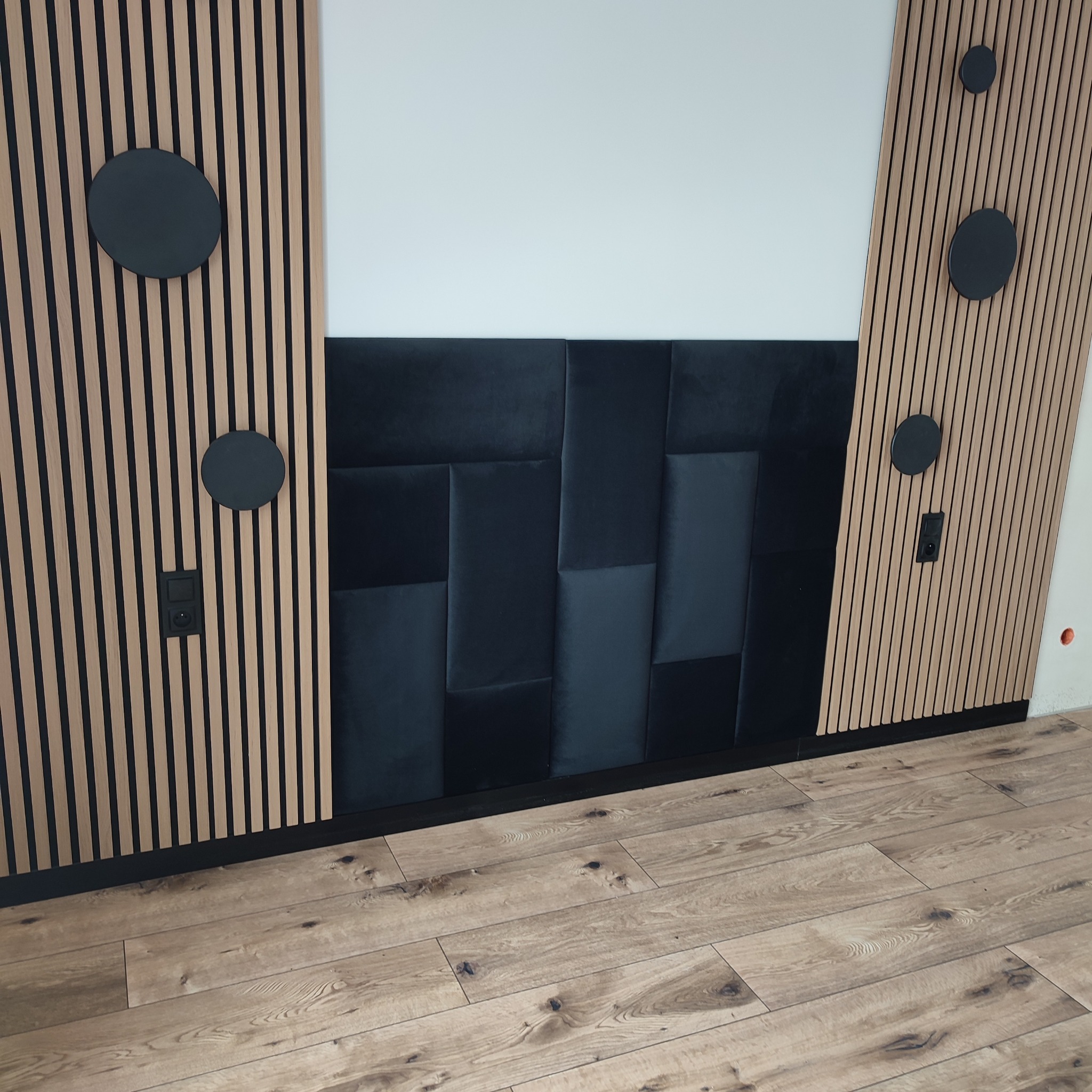 Nowoczesna aranżacja ściany z drewnianych lameli z czarnymi akcentami i tapicerowanym panelem. Widoczna podłoga z paneli w ciepłym odcieniu drewna.