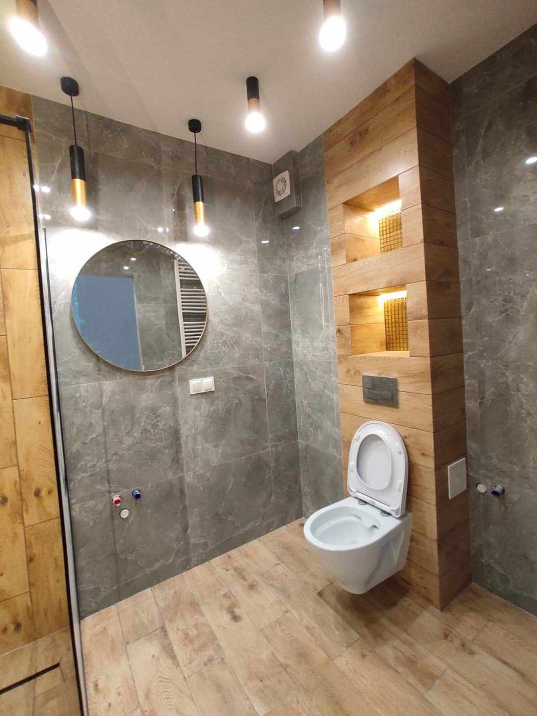 Nowoczesna łazienka z szarymi płytkami imitującymi marmur, drewnianą zabudową WC z wnękami i okrągłym lustrem. Podłoga wyłożona panelami imitującymi drewno.