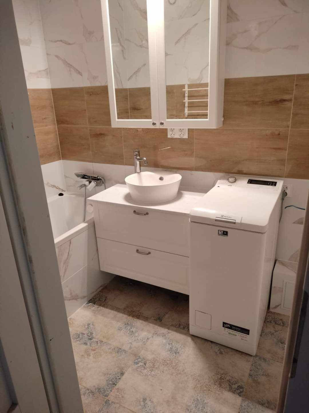 Jasna łazienka po remoncie: biała szafka z umywalką nablatową, pralka Electrolux, lustro z szafką, płytki imitujące marmur i drewno, wzorzysta podłoga.