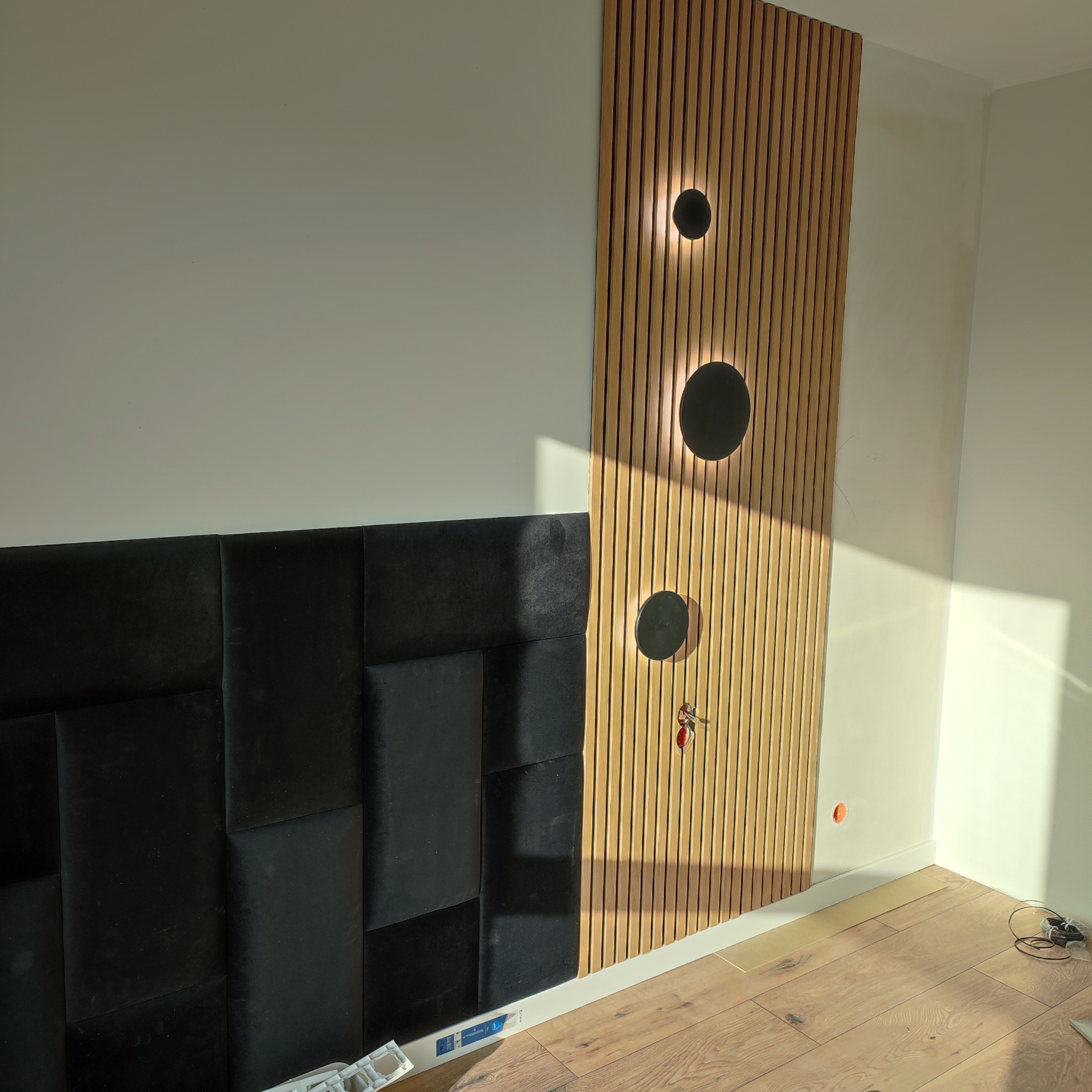 Panel z lameli drewnianych z okrągłymi lampami i czarny panel tapicerowany na ścianie w jasnym wnętrzu. Widoczne fragmenty podłogi z paneli.