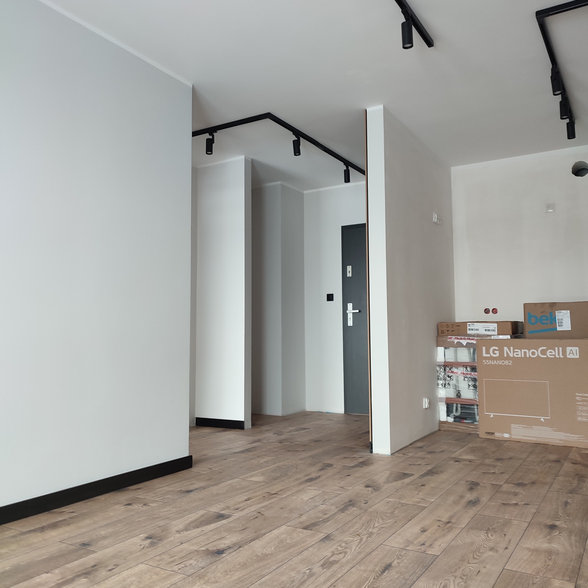 Jasne panele podłogowe w nowym mieszkaniu, widok na korytarz z czarnym oświetleniem szynowym i białe ściany. Kartony z nowym sprzętem RTV w tle.