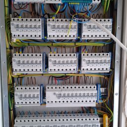 Elektron - Rozdzielnica elektryczna z zainstalowanymi bezpiecznikami, przewodami w różnych kolorach i elementami zabezpieczającymi. Widoczne detale montażu i podłączenia.