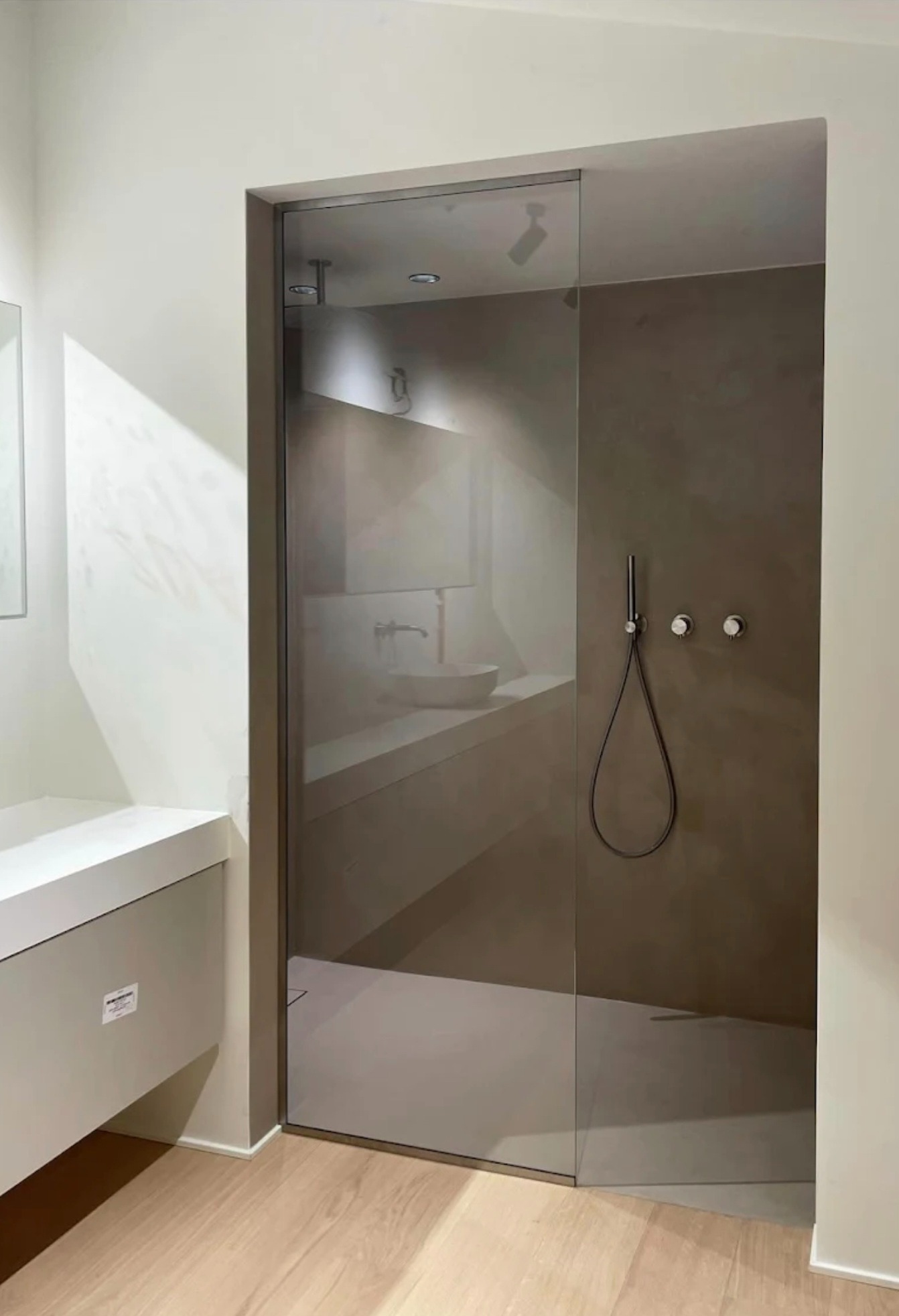 Minimalistyczna łazienka z kabiną prysznicową ze szkła hartowanego, widoczny deszczownica i bateria. Umywalka w tle. Jasne drewno na podłodze.