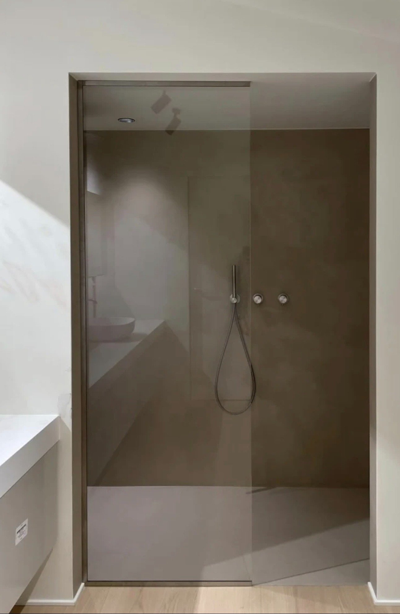 Minimalistyczna kabina prysznicowa ze szklanymi drzwiami przesuwnymi, z widoczną armaturą i elementami wystroju łazienki w stonowanej kolorystyce.